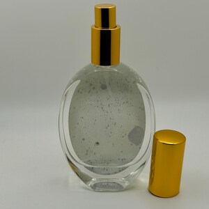 May include: Clear glass perfume bottle with a gold-colored cap and sprayer. The bottle has an oval shape and a gold-colored cap. The bottle is empty and has a speckled interior. The cap is off to the side.
