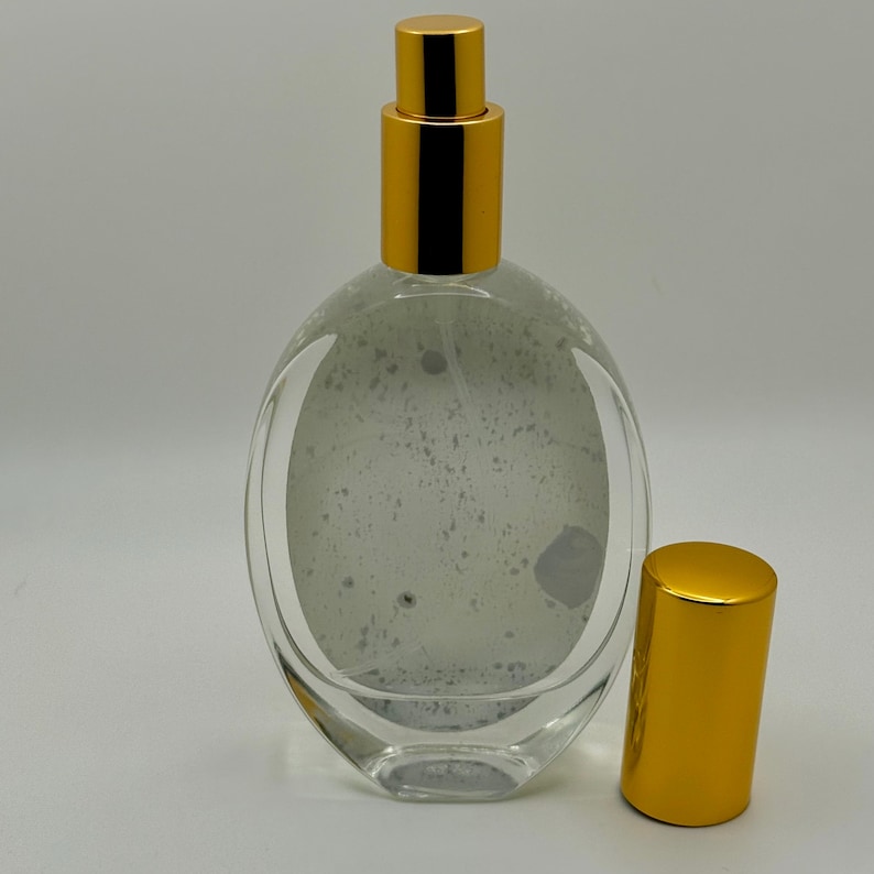 May include: Clear glass perfume bottle with a gold-colored cap and sprayer. The bottle has an oval shape and a gold-colored cap. The bottle is empty and has a speckled interior. The cap is off to the side.