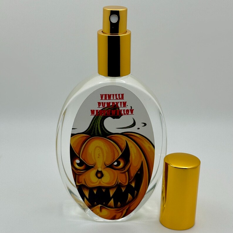 May include: A clear glass perfume bottle with a gold cap and sprayer. The label features a jack-o'-lantern design with the words "Vanilla Pumpkin Marshmallow". The bottle is oval-shaped and the cap is off to the side.
