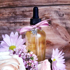 May include: A glass dropper bottle containing a golden liquid, decorated with a pink ribbon and a fairy charm. The bottle is surrounded by pink and purple flowers, including roses and daisies, on a wooden surface.