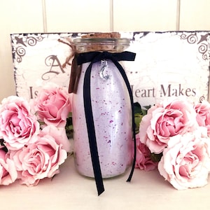 May include: A clear glass bottle filled with lavender-colored bath salts and rose petals, adorned with a black ribbon and a silver crescent moon charm. The bottle is topped with a cork and a small tag. Pink roses surround the bottle, with a decorative sign in the background.