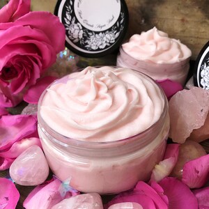 May include: A close-up of a pink whipped body butter in a glass jar. The jar is surrounded by pink rose petals and rose quartz crystals. The jar has a black lid with a white label that says "June Apothecary Los Angeles Rose Quartz Body Butter".