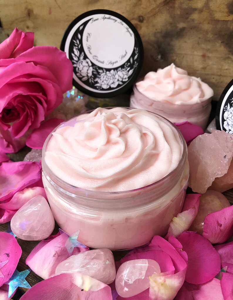 May include: A close-up of a pink whipped body butter in a glass jar. The jar is surrounded by pink rose petals and rose quartz crystals. The jar has a black lid with a white label that says "June Apothecary Los Angeles Rose Quartz Body Butter".