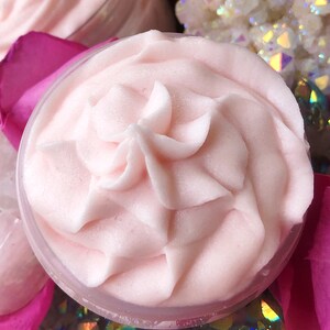 May include: Close-up of a pink whipped body butter in a clear jar. The butter is swirled and has a smooth, creamy texture.