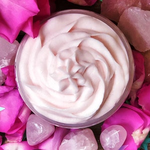 May include: A close-up of a pink whipped body butter in a clear glass jar. The jar is surrounded by pink rose petals and pink quartz crystals.