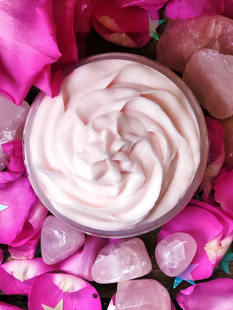 May include: A close-up of a pink whipped body butter in a clear glass jar. The jar is surrounded by pink rose petals and pink quartz crystals.