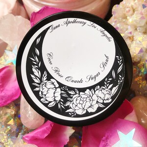 May include: A black jar with a white label that says "Luna Apothecary Los Angeles Cocoa Rose Quartz Sugar Scrub". The jar is surrounded by pink rose petals and crystals.