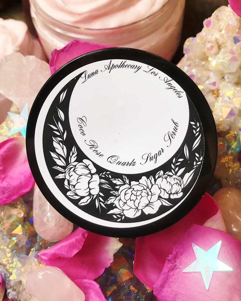 May include: A black jar with a white label that says "Luna Apothecary Los Angeles Cocoa Rose Quartz Sugar Scrub". The jar is surrounded by pink rose petals and crystals.