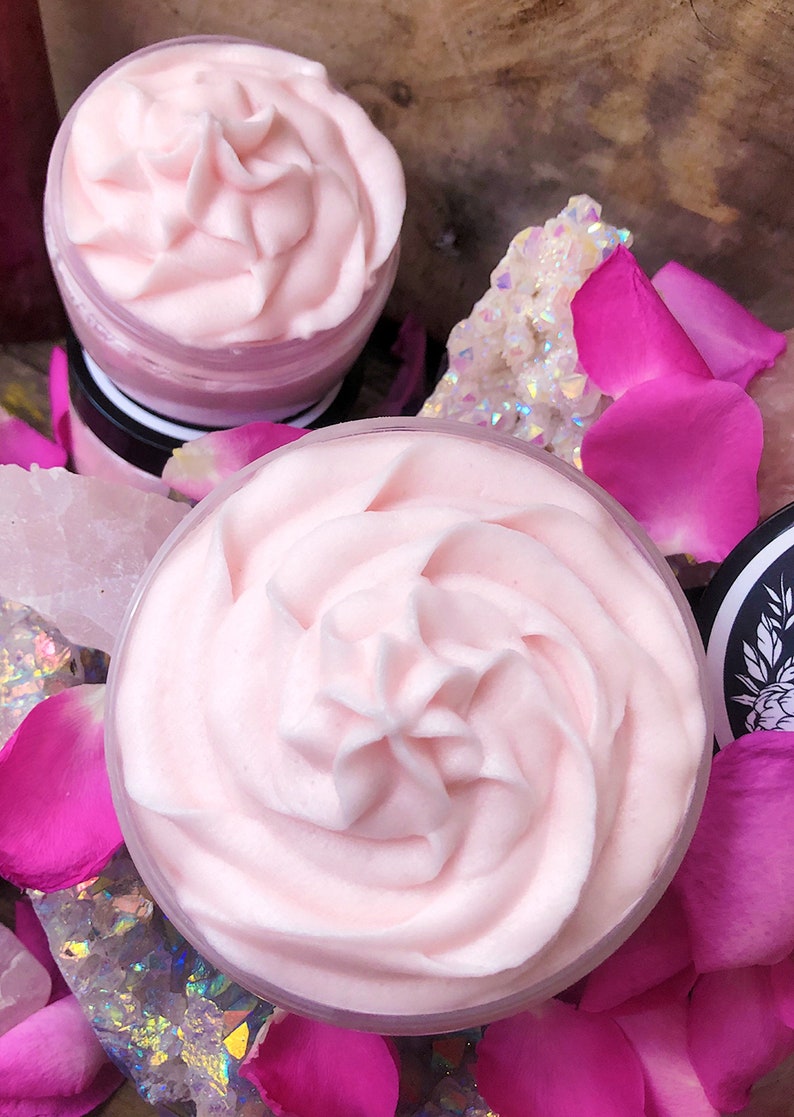 May include: Two containers of pink whipped body butter, one on top of the other, surrounded by pink rose petals and iridescent crystals.