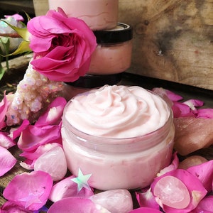 May include: A close-up of a pink whipped body butter in a glass jar. The jar is surrounded by pink rose petals and pink quartz crystals.