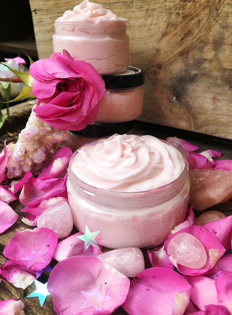 May include: A close-up of a pink whipped body butter in a glass jar. The jar is surrounded by pink rose petals and pink quartz crystals.
