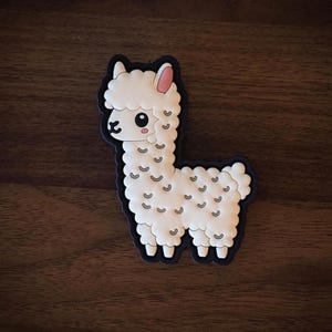 May include: A white llama-shaped sticker with a black outline. The llama has fluffy fur, pink inner ears, and small, curved markings on its body. The sticker is on a dark wood surface.