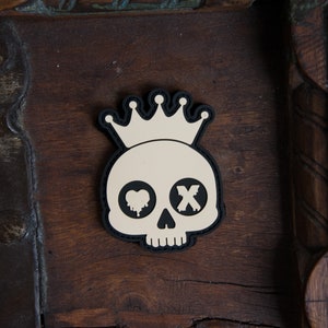 Puede incluir: Un parche de calavera blanco y negro con una corona en la parte superior. La calavera tiene un corazón para un ojo y una X para el otro ojo.