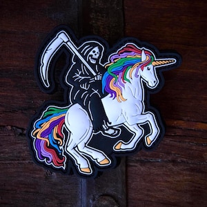 May include: A black patch featuring a Grim Reaper riding a white unicorn with a rainbow mane and tail. The Reaper is holding a scythe. The patch has a black border and is set against a wooden background.
