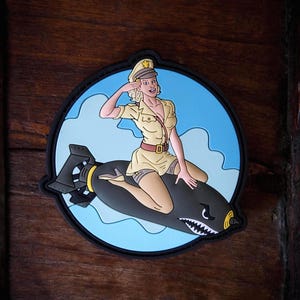 Puede incluir: Un parche circular con una imagen de dibujos animados de una mujer con uniforme militar saludando mientras monta una bomba. La bomba es negra con un diseño de boca de tiburón blanco. El fondo es un cielo azul con nubes blancas.