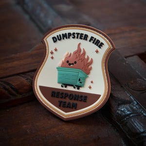 May include: A shield-shaped patch with the words "DUMPSTER FIRE" and "RESPONSE TEAM". The patch features a cartoon dumpster on fire with a smiling face. The dumpster is teal, the flames are brown, and the background is off-white.
