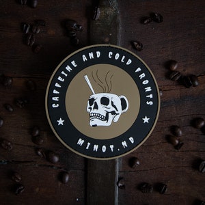 Puede incluir: Un parche redondo negro y marrón con el texto "Caffeine and Cold Fronts" y una calavera en una taza de café con vapor saliendo de ella. El texto "Minot, ND" está en la parte inferior del parche.