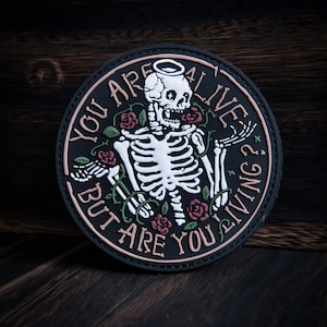 May include: Black and white patch with a skeleton with a halo, surrounded by roses and vines. The text reads "You are alive but are you living?"