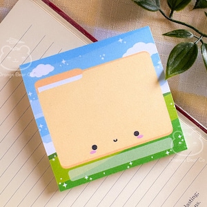 May include: A square sticky note with a cartoon file folder design. The file folder is light yellow with a smiling face, set against a blue sky and green grass background. The note is on a lined notebook page.