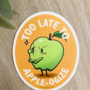May include: A round sticker with an orange background and a green apple with a face. The apple is frowning and has its arms crossed. The text "Too Late To Apple-O-gize" is written around the apple.