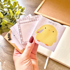 May include: A set of four square sticky notes with different designs. One note features a white ghost with the word "BOO!" on it. Another note has a gray background with a white checkered border and a blank space for writing. A third note has a grid pattern and a yellow chick holding a knife with the text "JUST DO IT". The final note has a white background with a grid pattern.