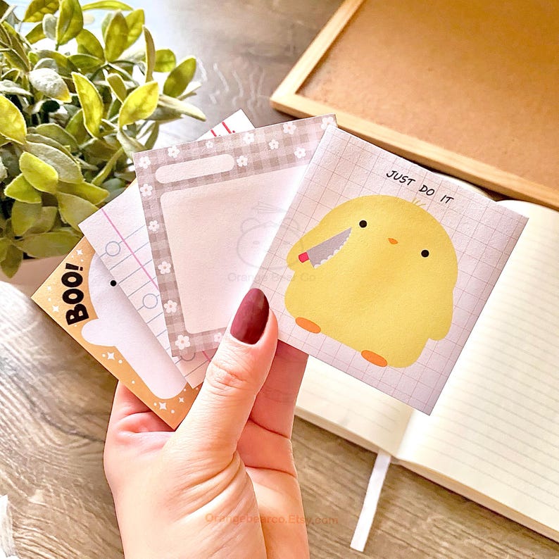 May include: A set of four square sticky notes with different designs. One note features a white ghost with the word "BOO!" on it. Another note has a gray background with a white checkered border and a blank space for writing. A third note has a grid pattern and a yellow chick holding a knife with the text "JUST DO IT". The final note has a white background with a grid pattern.