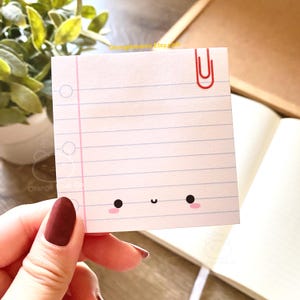 May include: A white lined notepad with a cute cartoon face and a red paperclip. The notepad has pink lines and a white background.