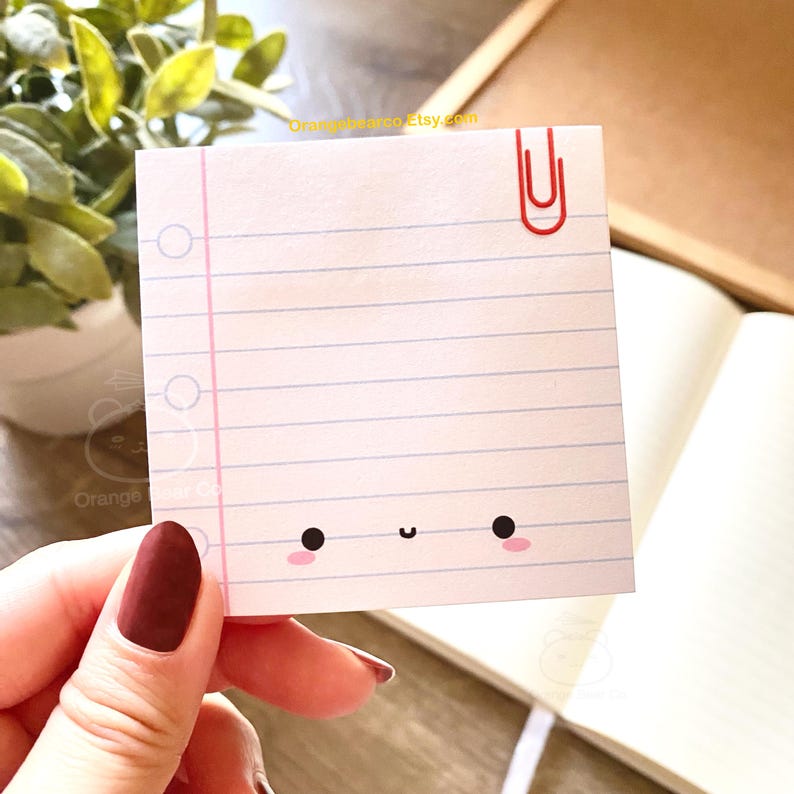 May include: A white lined notepad with a cute cartoon face and a red paperclip. The notepad has pink lines and a white background.