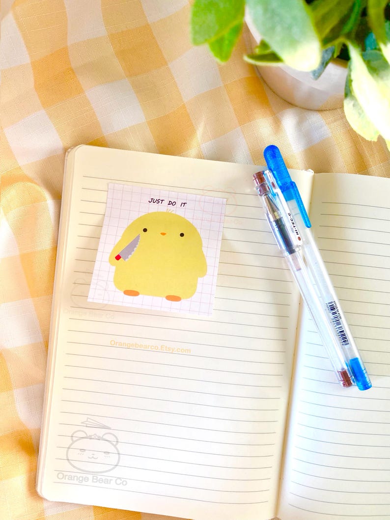 May include: An open notebook with lined pages and a yellow sticky note with a cartoon chicken holding a knife and the text "Just Do It". The notebook is on a yellow and white checkered surface. Two pens are on the right side of the notebook.