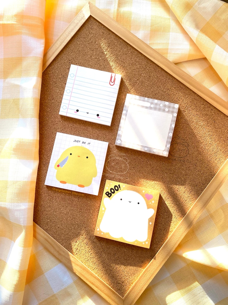 May include: Four different sticky note pads with cute illustrations. The note pads are on a cork board with a yellow and white checkered fabric in the background. The note pads feature a ghost with the text "BOO!" a chicken with a knife and the text "Just do it", a lined note pad with a cute face, and a blank note pad with a gray and white patterned border.