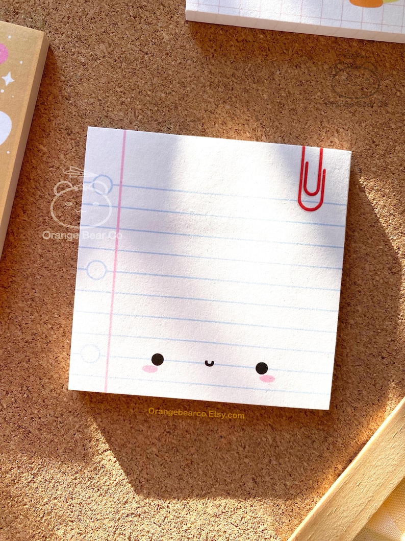 May include: A white notepad with blue lines and a pink border. The notepad has a cute cartoon face with black eyes and a pink blush. A red paperclip is attached to the top right corner. The notepad has the text "Orangebearco Etsy.com" at the bottom.