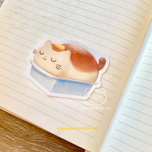 Puede incluir: Una pegatina con un gato de dibujos animados durmiendo dentro de una hogaza de pan con una porción de mantequilla en la parte superior. La pegatina es blanca con un fondo azul y tiene el texto "Orange Bear Co" en la parte inferior.