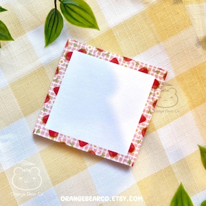 May include: A square notepad with a white center and a strawberry-themed border. The border features a pink and white gingham pattern with red strawberry illustrations. The notepad is on a yellow and white checkered surface.