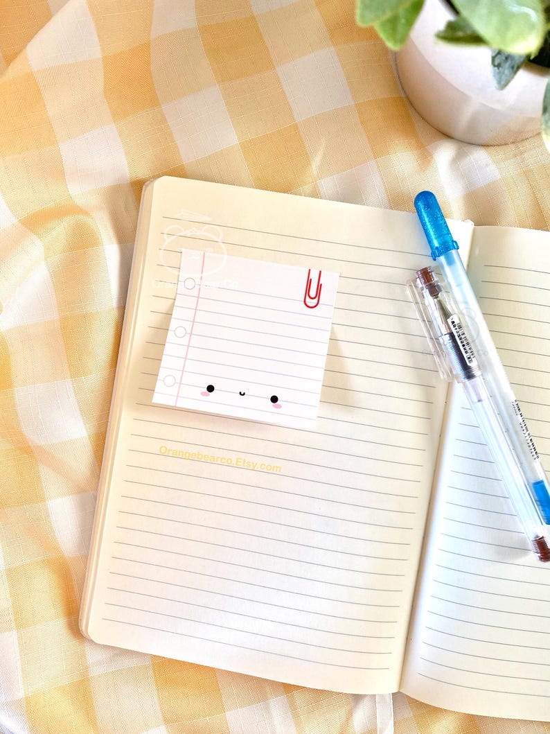 May include: A white lined notepad with a cute cartoon face and a red paperclip attached to the top. The notepad is sitting on a yellow and white checkered surface with a blue pen and a potted plant in the background.