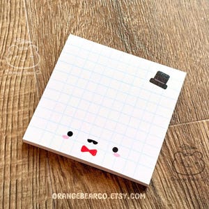 May include: A white notepad with a grid pattern and a cute cartoon face with a red bow tie and a black top hat. The notepad has the text "ORANGEBEARCO.ETSY.COM" printed on the bottom.