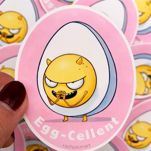 Puede incluir: Una pegatina con un personaje de huevo de dibujos animados con una expresión traviesa. El huevo es amarillo con una cáscara blanca y azul, sobre un fondo rosa. El texto "Egg-Cellent" se muestra debajo.