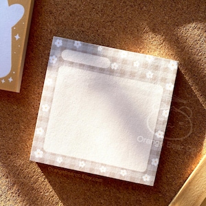 Może przedstawiać: Biały notes z obramowaniem w kratkę i małymi białymi kwiatami. Notes ma pustą przestrzeń do pisania z cienką białą linią u góry.