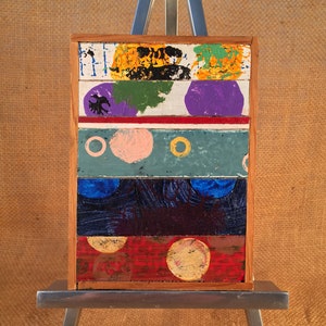 May include: A colorful abstract painting on wood, featuring geometric shapes and circles in shades of yellow, green, purple, pink, blue, and red. The painting is displayed on a small easel.