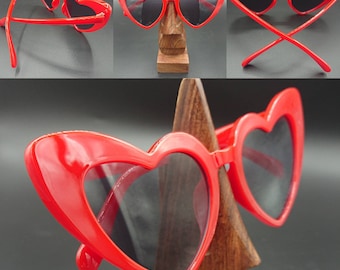 May include: Red heart-shaped sunglasses with black lenses. The sunglasses are on a wooden stand.