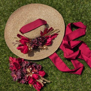 May include: A wide-brimmed, gold-colored sun hat with a burgundy ribbon and floral embellishments. A matching burgundy ribbon and a separate floral arrangement are also present, all set against a green grass background. The hat is a stylish accessory.