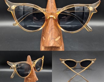 May include: A pair of cat-eye sunglasses with a clear amber frame and black lenses. The sunglasses are displayed on a wooden stand.