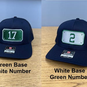 May include: Two navy blue baseball caps with mesh backs and adjustable snapback closures. One cap features a green rectangular patch with the white number 17, while the other displays a white rectangular patch with the green number 2. Both have stickers.