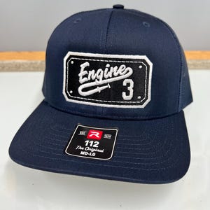 May include: A navy blue baseball cap with a mesh back and a flat brim. A rectangular black patch on the front features white embroidered text that reads Engine 3. A black sticker on the brim displays the text Est. 1970 R 112 The Original MD-LG.