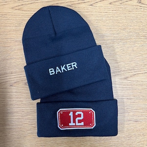 May include: Two navy blue knit beanies stacked on top of each other. The top beanie features the word BAKER embroidered in white thread. The bottom beanie displays a rectangular red patch with white stitching and the white number 12 in the center.