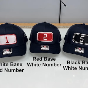 May include: Three navy blue trucker caps with mesh backs and adjustable snapback closures. Each cap features a rectangular patch on the front with a number: a white patch with a red number 1, a red patch with a white number 2, and a black patch with a white number 5.