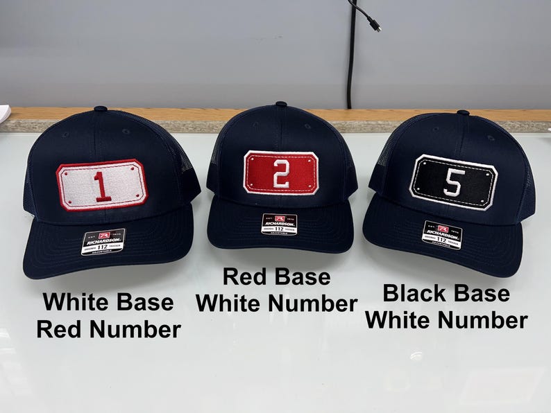 May include: Three navy blue trucker caps with mesh backs and adjustable snapback closures. Each cap features a rectangular patch on the front with a number: a white patch with a red number 1, a red patch with a white number 2, and a black patch with a white number 5.