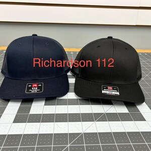 May include: Two Richardson 112 trucker hats, one in navy blue and one in black, featuring a structured front panel, a mesh back, and a flat brim. Each hat displays a rectangular sticker on the brim with the text Richardson 112 The Original.