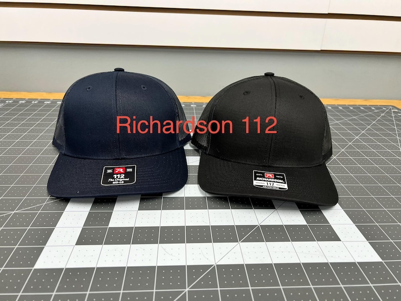 May include: Two Richardson 112 trucker hats, one in navy blue and one in black, featuring a structured front panel, a mesh back, and a flat brim. Each hat displays a rectangular sticker on the brim with the text Richardson 112 The Original.
