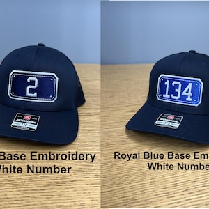 May include: Two baseball caps with mesh backs and adjustable straps. The left cap is navy blue with a white embroidered number 2 on a navy patch. The right cap is navy blue with a white embroidered number 134 on a royal blue patch. Both have stickers.