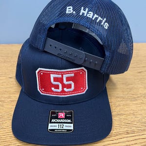 May include: A navy blue trucker hat with a mesh back and a snapback closure. The front features a rectangular red patch with the white number 55. The back mesh panel is embroidered with the white text B. Harris. A Richardson 112 sticker is on the brim.